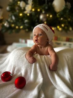 a baby laying on top of a white blanket next to a christmas tree with ornaments