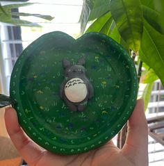 a hand holding a green plate with a cartoon animal on it's face in front of a potted plant