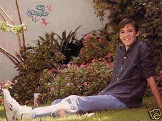 a young boy sitting on the grass in front of some bushes and flowers smiling at the camera