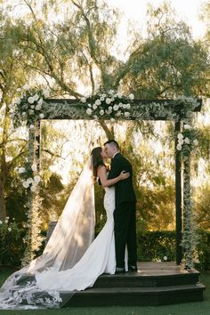 a bride and groom kissing in front of an outdoor wedding ceremony arch with white flowers