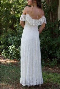 a woman wearing a white dress standing in front of some trees and bushes with her back to the camera