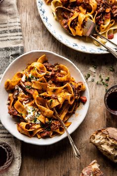 two plates of pasta with meat and cheese on them next to some bread, wine glasses and utensils