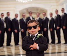 a young boy wearing sunglasses and standing in front of a group of men with their arms crossed