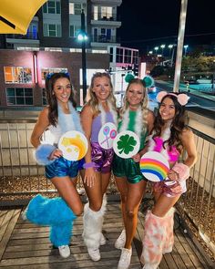 four women dressed in costume posing for a photo on a boardwalk at night with one holding a plate