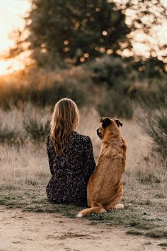 a woman sitting on the ground next to her dog looking up at the sky with trees in the background