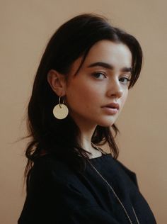 a woman with long dark hair wearing a black top and gold earrings, standing in front of a beige wall