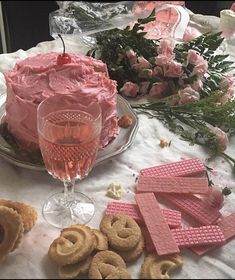 a table topped with cookies and pink frosted cake next to wine glasses filled with flowers