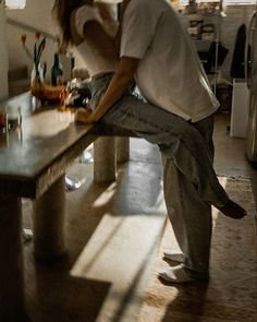 a man sitting on top of a kitchen counter next to a wooden table covered in bottles