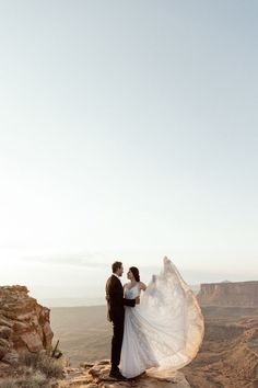 a bride and groom standing on the edge of a cliff at sunset with their veil blowing in the wind