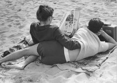 two people laying on the sand reading a book