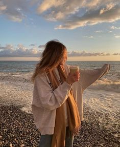 a woman standing on top of a beach next to the ocean holding a coffee cup