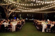 a large group of people are sitting at tables under string lights in the evening time