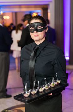 a woman in a black mask and tie holding a tray with glasses filled with drinks