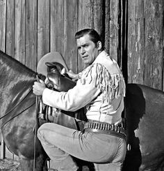 a man kneeling next to a horse in front of a wooden fence with a cowboy hat on