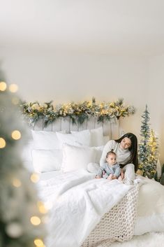 a mother and her baby sitting on a bed with christmas lights in the corner behind them