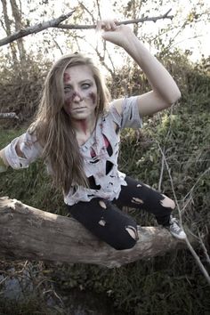 a woman with makeup on her face sitting on a tree branch in the woods posing for a photo