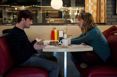 a man and woman sitting at a table with food in front of them, talking