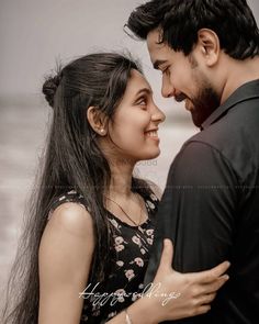 a man and woman standing next to each other in front of the ocean with their arms around each other