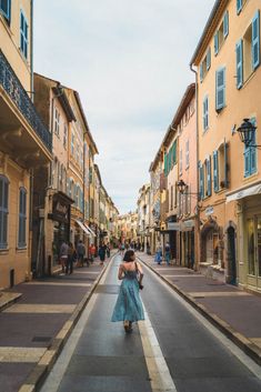 a woman in a blue dress walking down the middle of an empty street with buildings on both sides