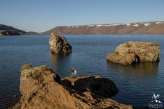 two people standing on the edge of a large body of water with mountains in the background