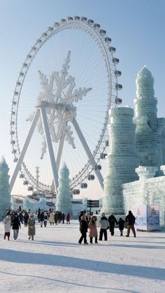 people are walking around an ice sculpture in the middle of winter with a large ferris wheel behind them