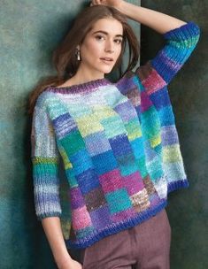 a woman wearing a multicolored sweater leaning against a wall with her hands on her head