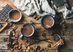 two mugs of hot chocolate on a wooden table next to cinnamon sticks and spices