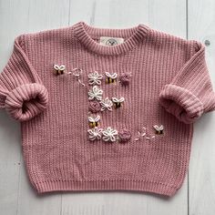 a pink sweater with flowers and butterflies on the front is laying on a white wooden surface