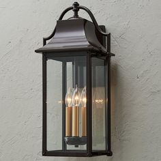 an outdoor light with three lit candles on the side of it, against a white wall