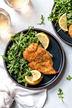 two plates filled with fish and greens on top of a white table next to glasses