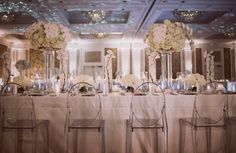 the tables are set up with clear chairs and white flowers in vases on them