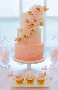 a three tiered cake with pink flowers on top and cupcakes in the foreground