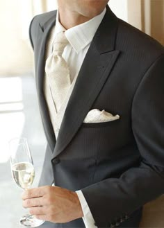 a man in a tuxedo holding a glass of wine and looking at the camera