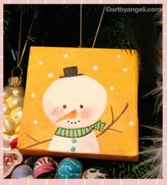 an ornament hanging from a christmas tree with ornaments around it and a painting of a snowman