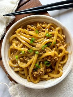 a white bowl filled with noodles and meat on top of a wooden cutting board next to chopsticks