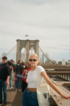 a woman standing on top of a bridge holding onto a white bag with the brooklyn bridge in the background