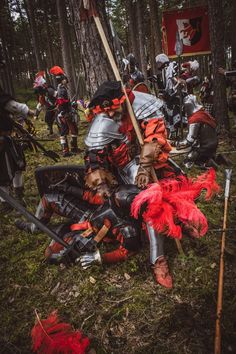 some people dressed in medieval armor and red feathers are sitting on the ground near trees