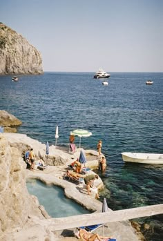 some people are sitting on the rocks by the water and boats in the water behind them