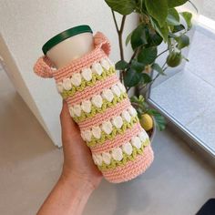 a hand holding a coffee cup in a crocheted case next to a potted plant