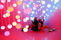 a man and woman sitting on the ground in front of a pink wall with lights hanging from it