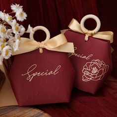 two red gift bags with gold lettering on them and white flowers in the vase behind them