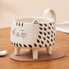 a ceramic cat shaped cup sitting on top of a wooden table next to a spoon