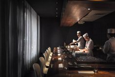 two chefs preparing food in a restaurant setting with long wooden tables and chairs around them