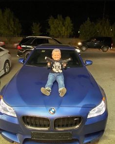 a baby doll is sitting on the hood of a blue car in a parking lot