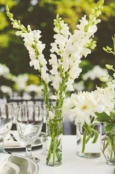 white flowers are in vases on a table set with silverware and place settings