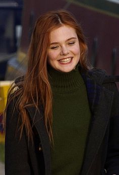 a woman with red hair is smiling and wearing a green turtle - neck sweater while walking down the street