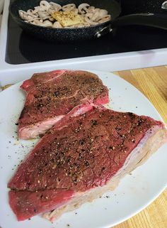 two steaks on a white plate next to a frying pan with mushrooms and seasonings