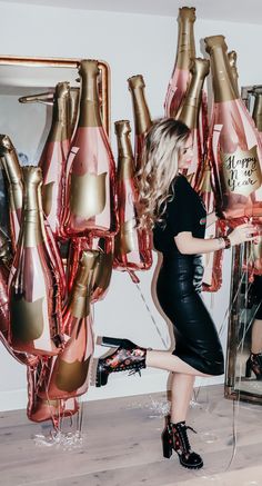 a woman in a black dress is standing near some pink and gold balloons that look like champagne bottles