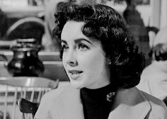 an old photo of a woman sitting at a table in front of some chairs and looking off to the side