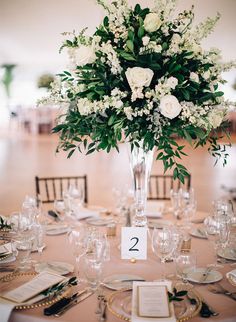 a tall vase filled with white flowers and greenery on top of a round table